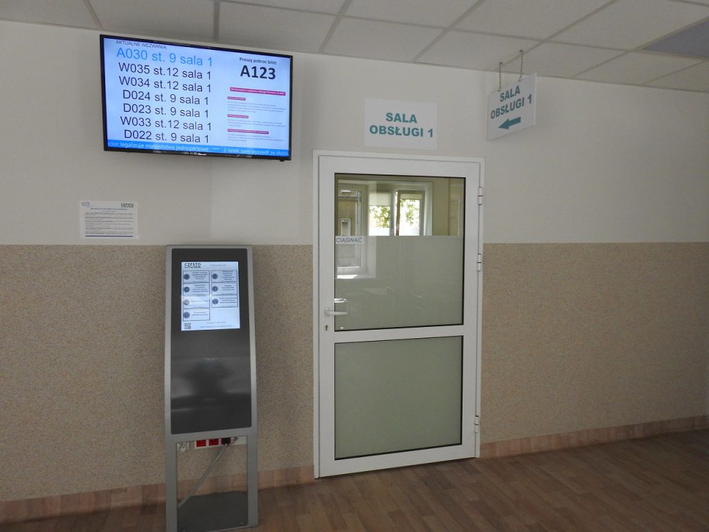 Drzwi prowadzące do sali obsługi numer 1 przed, którymi znajduje się biletomat systemu kolejkowego oraz ekran wyświetlający numery kolejki.