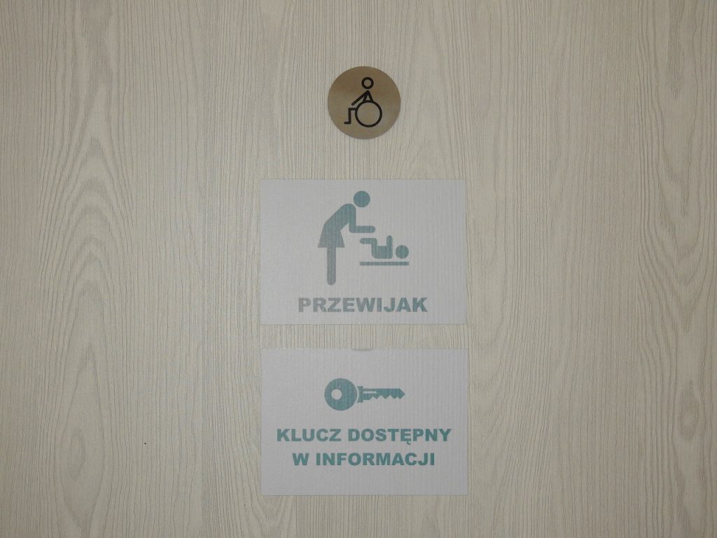 Oznaczenie na drzwiach toalety dla niepełnosprawnych z informacją, że klucz dostępny jest w informacji.